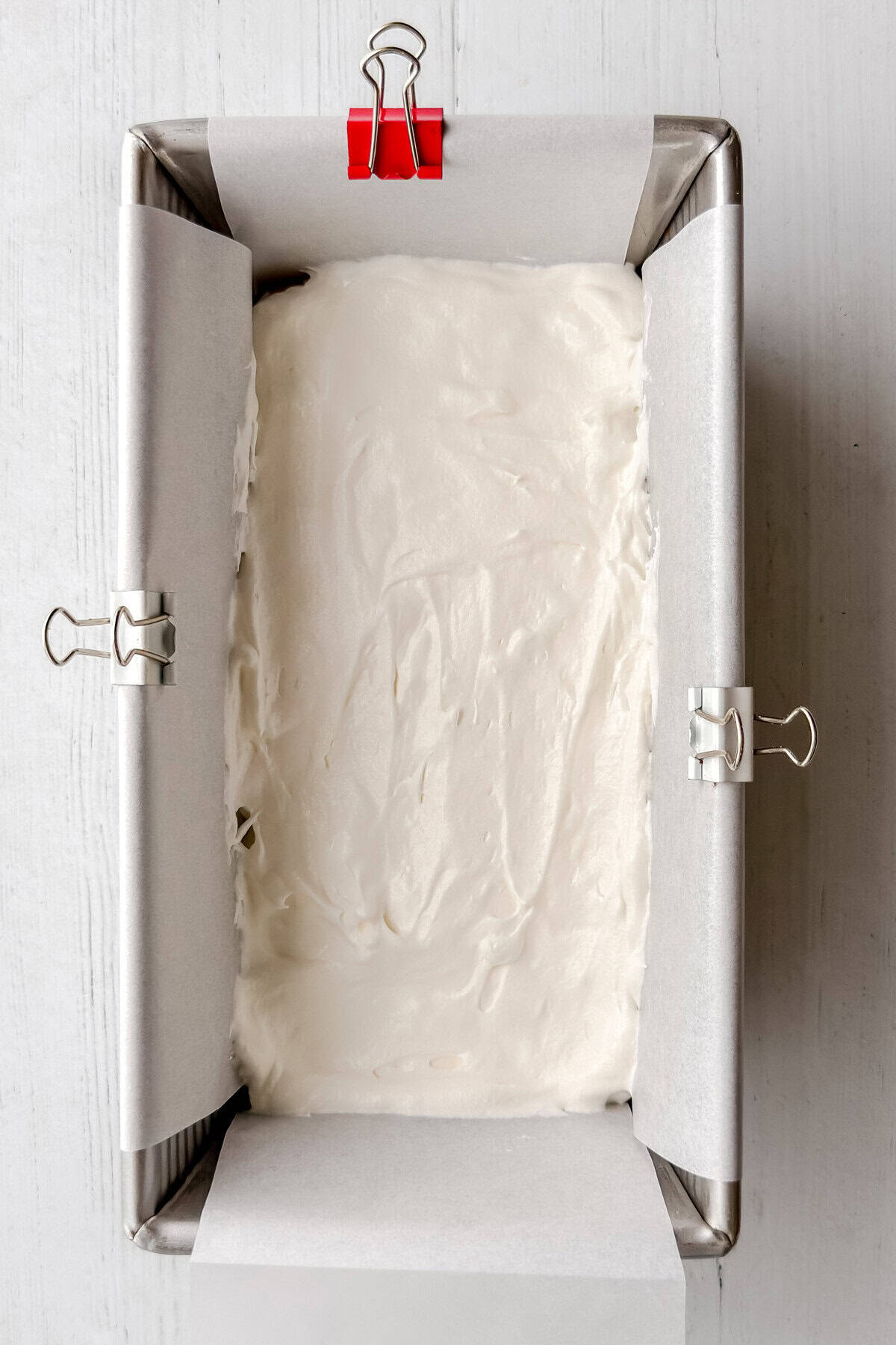 A metal baking pan lined with parchment paper, secured by binder clips, holds a layer of smooth white batter for a no-bake s'mores icebox cake, all set on a white surface.