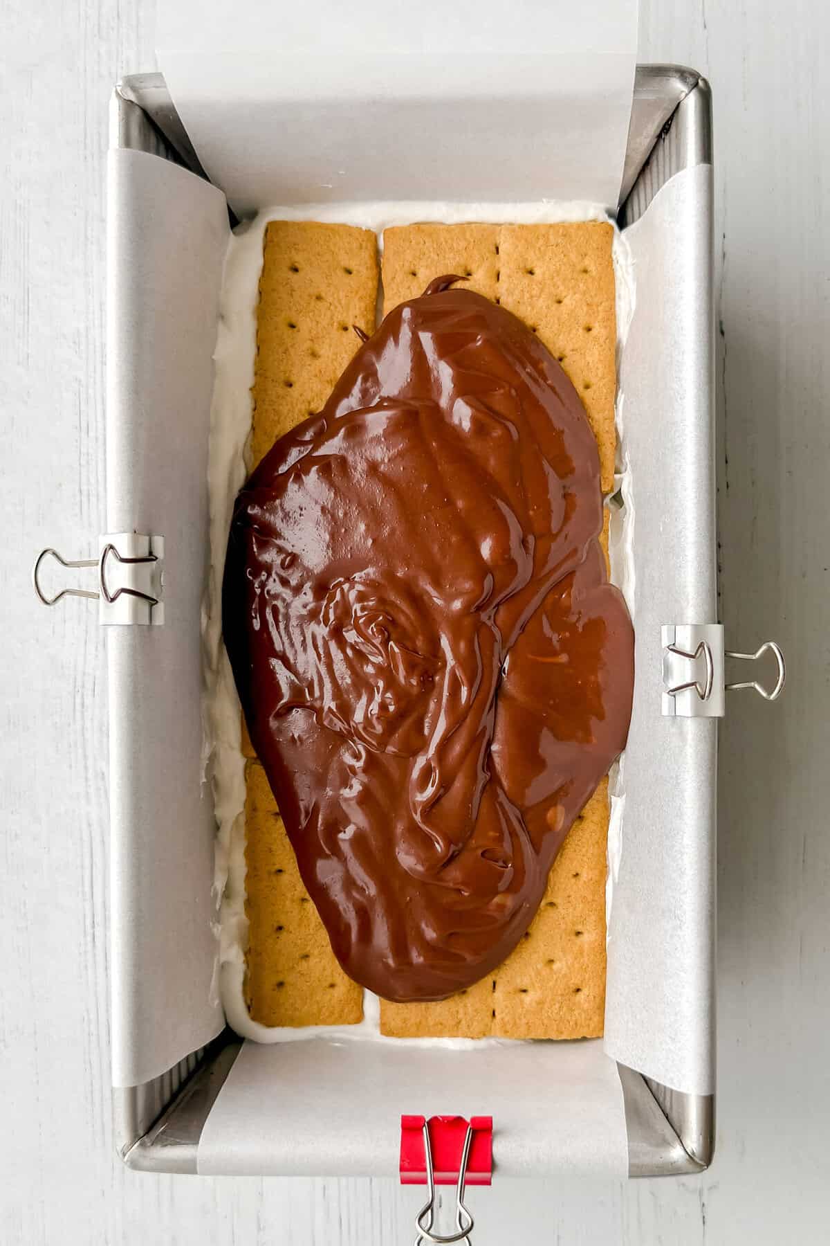 A loaf pan lined with parchment paper and binder clips holds a layer of graham crackers, topped with a thick layer of chocolate ganache for an irresistible no-bake s'mores icebox cake.