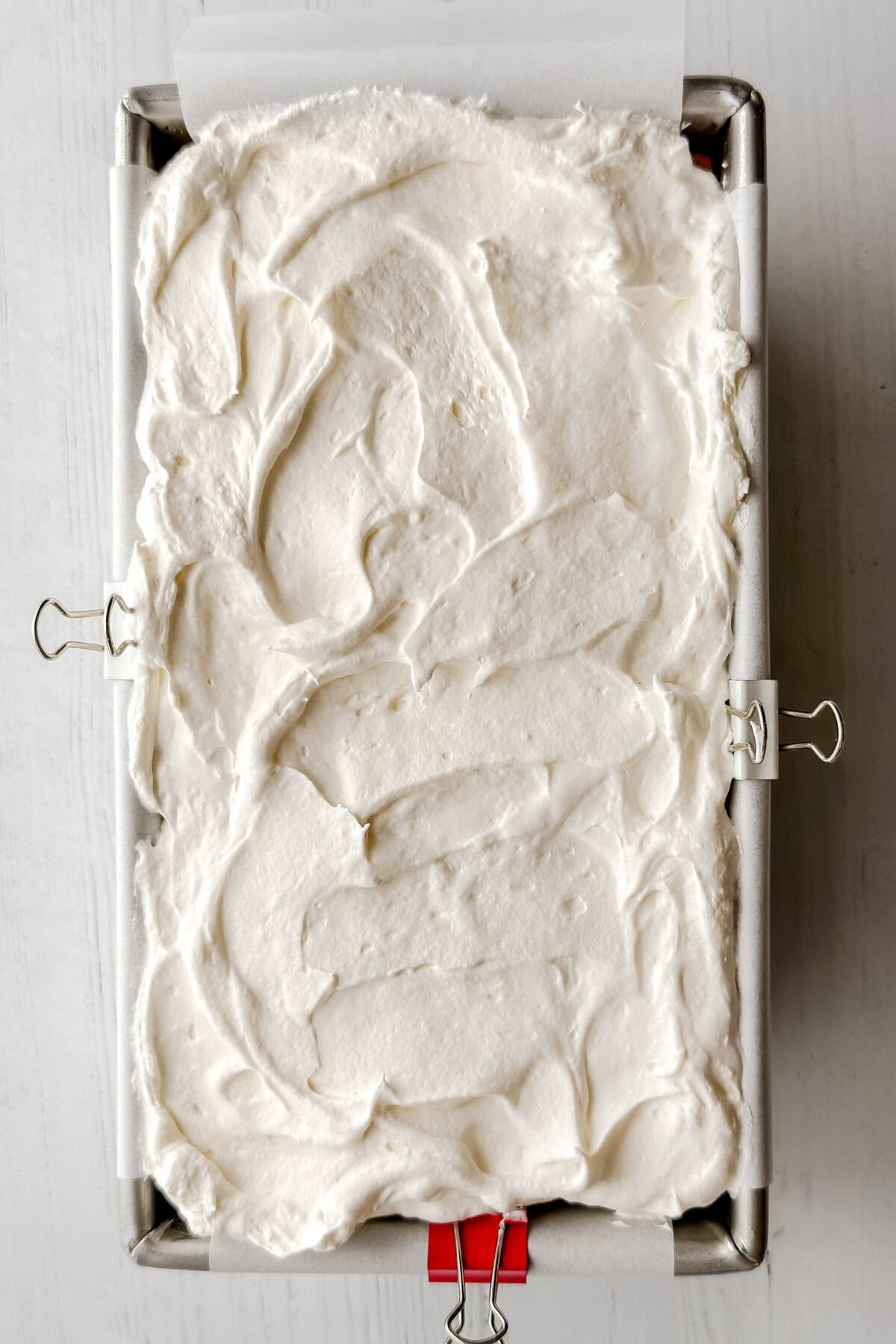 A rectangular metal pan lined with parchment paper, held in place by binder clips, filled with a smooth, swirled layer of white whipped cream—perfect for assembling a no-bake s'mores icebox cake.