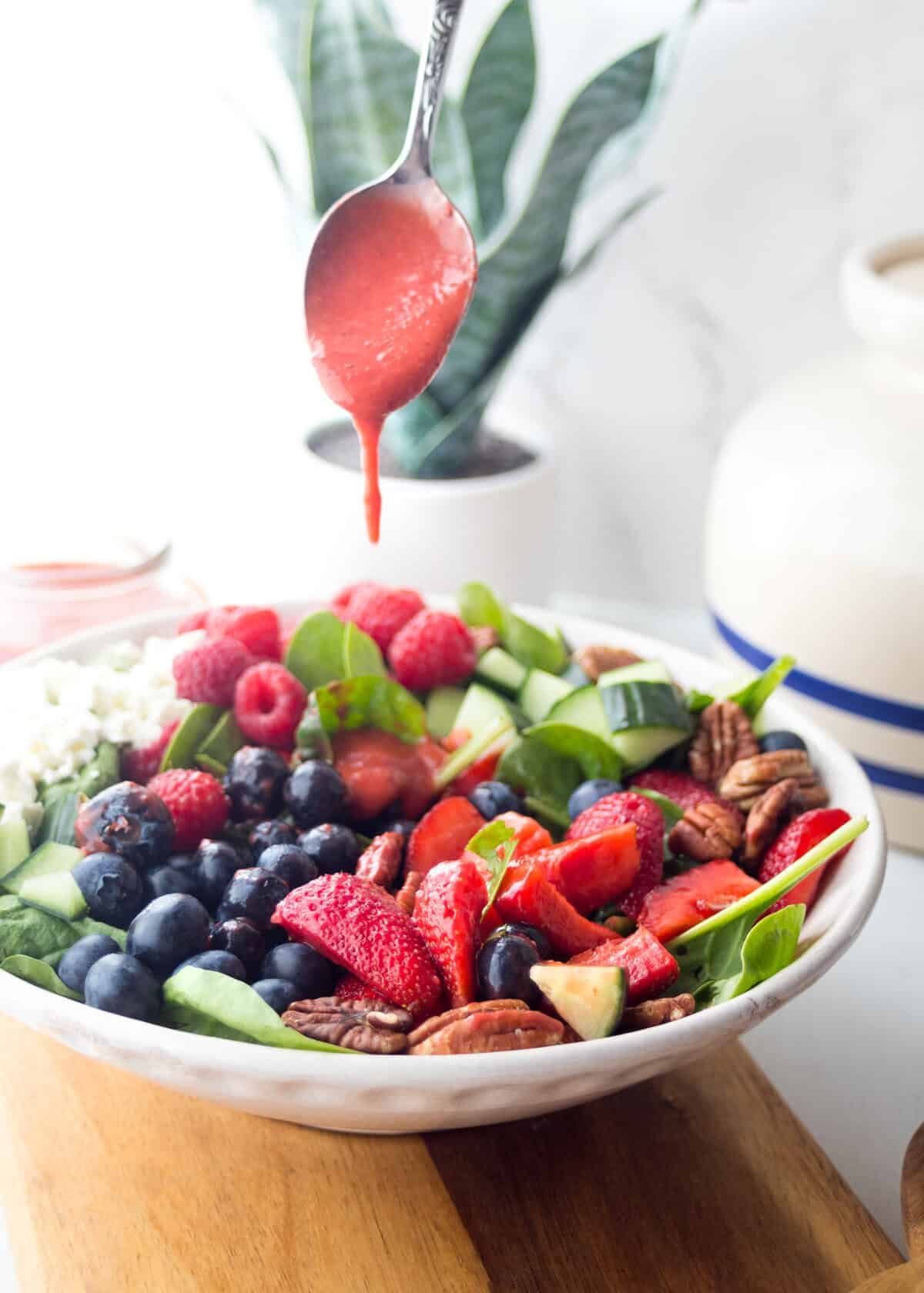 A vibrant spinach strawberry salad with blueberries, raspberries, cucumber, pecans, and feta fills a white bowl as strawberry dressing is drizzled from above. In the background, a houseplant and ceramic jar add a cozy touch.