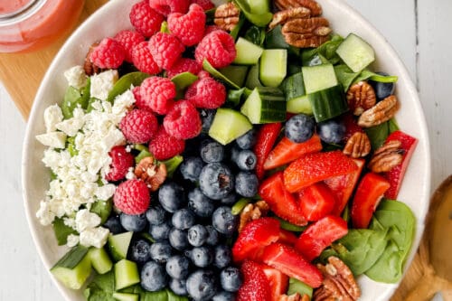 A spinach strawberry salad bowl with fresh blueberries, raspberries, cucumber, pecans, and crumbled cheese sits on a wooden board beside a jar of pink dressing and a checkered napkin.