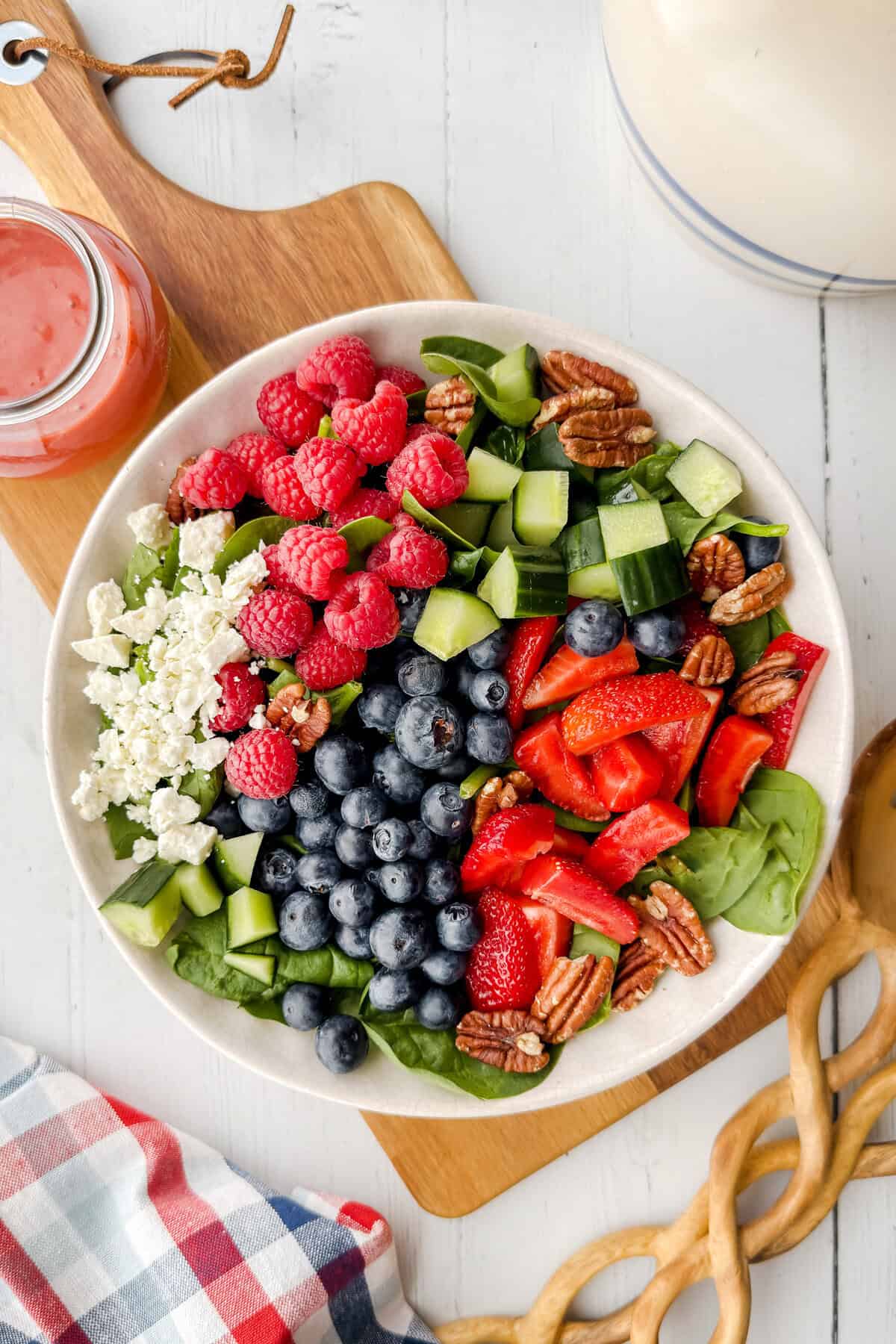A spinach strawberry salad bowl with fresh blueberries, raspberries, cucumber, pecans, and crumbled cheese sits on a wooden board beside a jar of pink dressing and a checkered napkin.