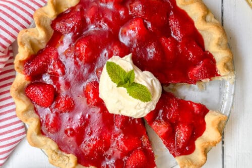 A strawberry pie recipe with a golden crust, topped with whipped cream and mint, sits on a white table. One slice is cut. A gold pie server, striped napkin, and floral d&eacute;cor are nearby.