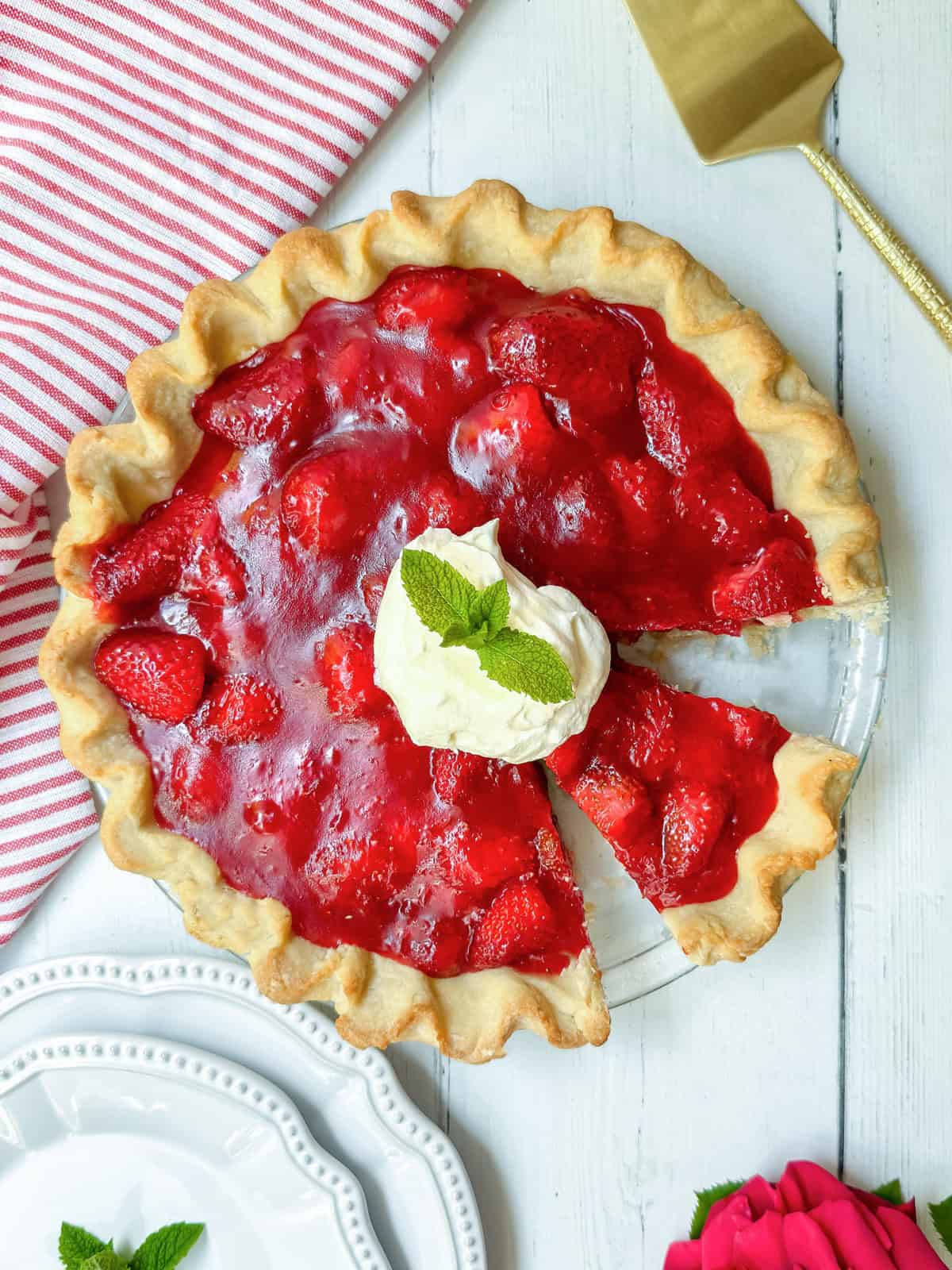 A strawberry pie made from a classic strawberry pie recipe, with a slice cut out, is topped with whipped cream and a mint sprig. It sits on a white wooden table next to a pie server and striped napkin.