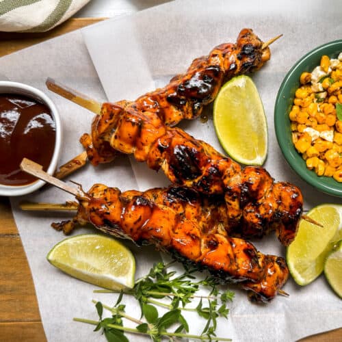 Three grilled chicken skewers on parchment paper with lime wedges, a bowl of barbecue sauce, and a bowl of seasoned corn on a wooden tray, garnished with fresh herbs.