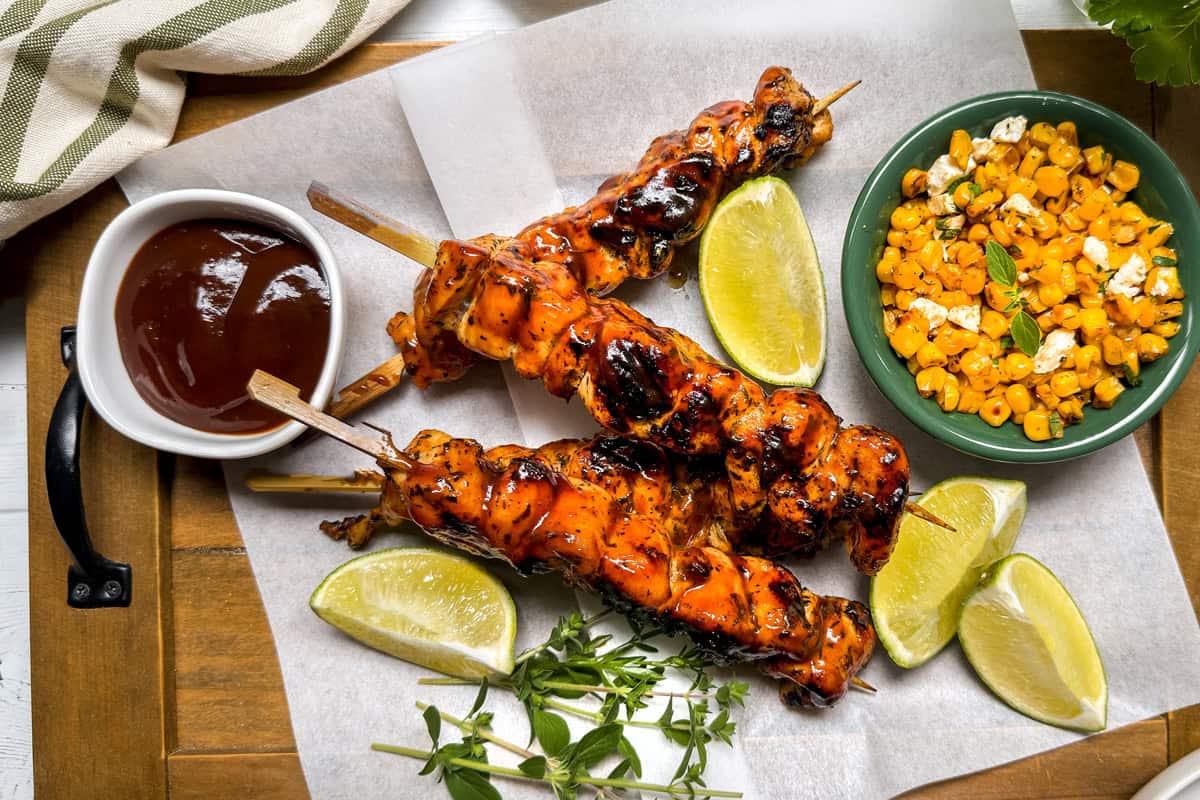 Three grilled chicken skewers on parchment paper with lime wedges, a bowl of barbecue sauce, and a bowl of seasoned corn on a wooden tray, garnished with fresh herbs.