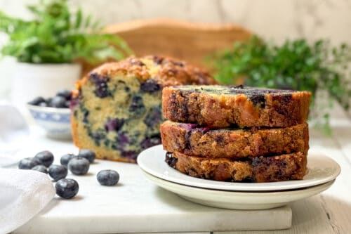 Three slices of blueberry muffin bread are stacked on a white plate in front of a loaf. Fresh blueberries are scattered on the table, with green plants in the background.