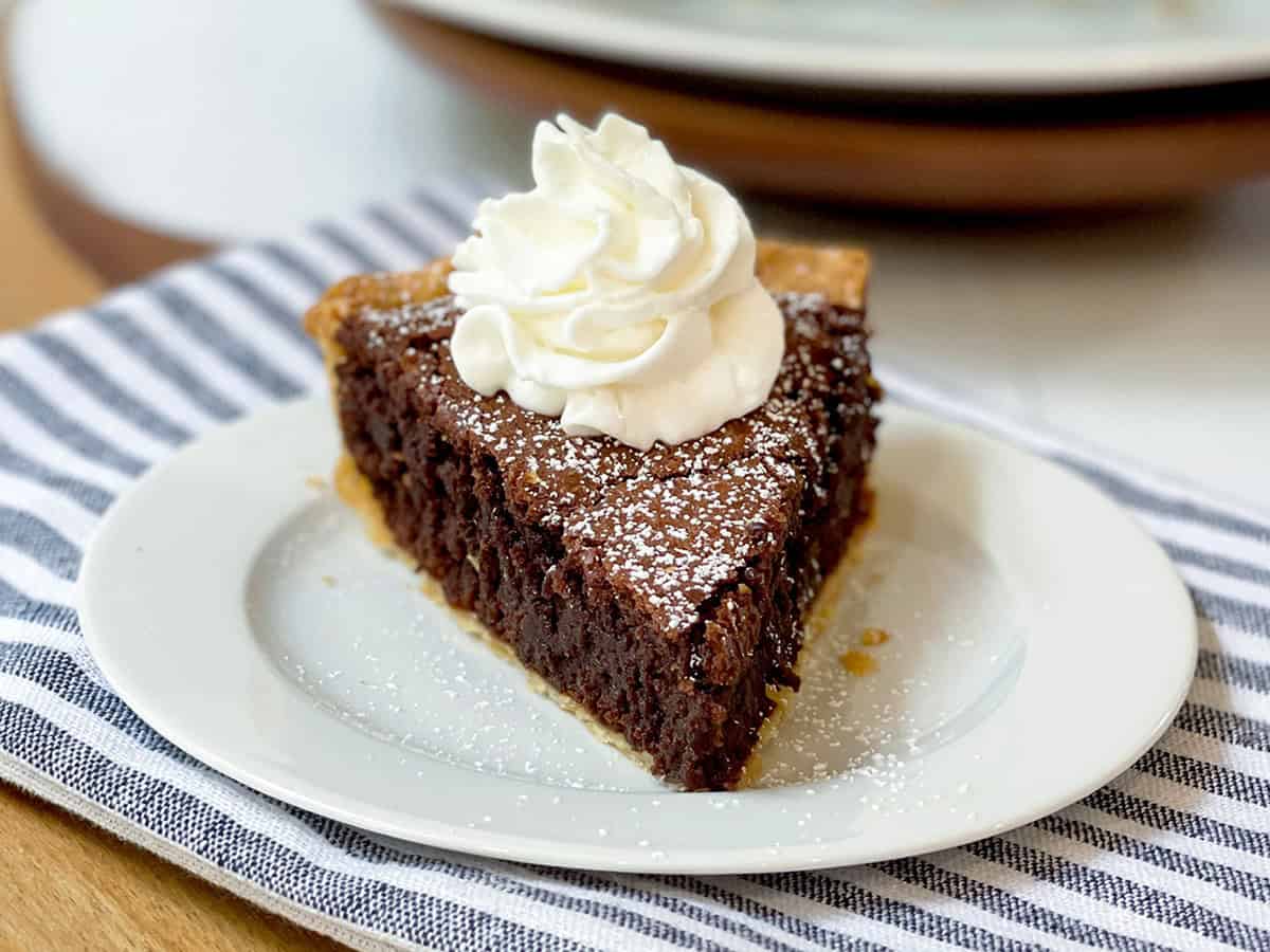 A slice of chocolate pie topped with whipped cream and powdered sugar on a white plate, placed on a striped cloth-perfect for Father's Day Dessert Recipes.
