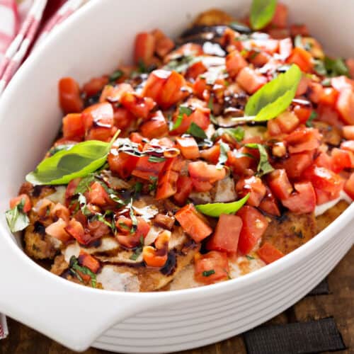 A white baking dish filled with baked chicken breasts topped with diced tomatoes, fresh basil leaves, and a drizzle of balsamic glaze, set on a wooden board next to a red and white checkered napkin.