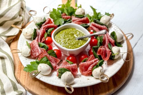 A white plate displays skewers with salami, mozzarella balls, basil, and cherry tomatoes arranged in a ring around a bowl of green pesto dip, garnished with fresh herbs. A spoon rests in the dip.