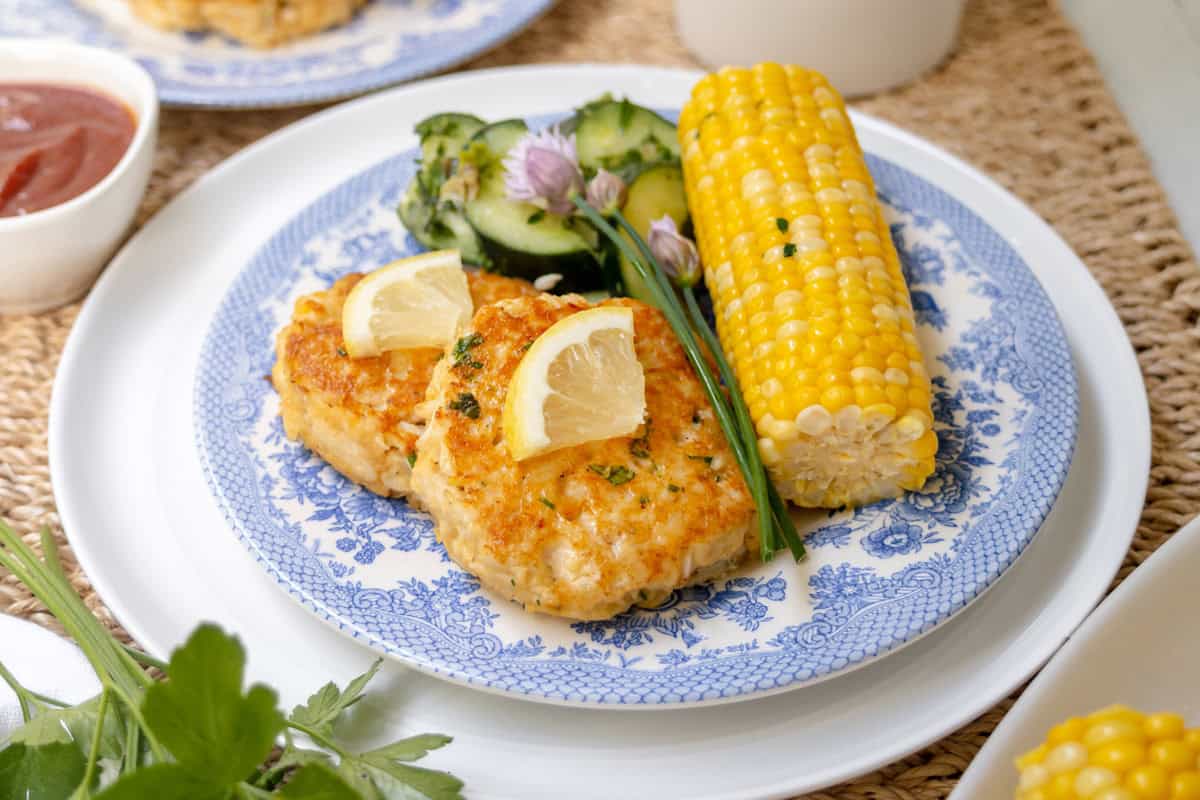 A plate with two Maryland crab cakes garnished with lemon wedges, a serving of cucumber salad, and a cob of corn on the blue and white plate. A small bowl of sauce is visible in the background.