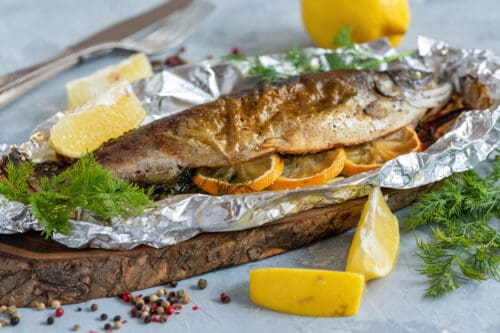 A whole baked fish with lemon slices and herbs, served in foil on a wooden board, garnished with fresh dill, lemon wedges, and peppercorns, with a fork and knife in the background.