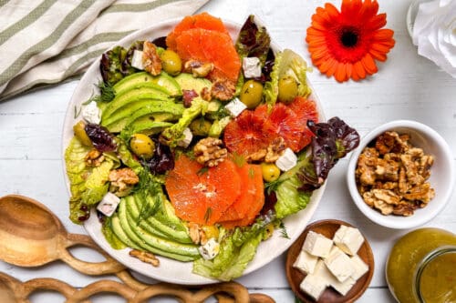 A colorful salad with avocado slices, orange and blood orange rounds, lettuce, olives, walnuts, and feta on a white plate. A wooden spoon, feta cubes, walnuts, and a bright orange flower surround the plate.