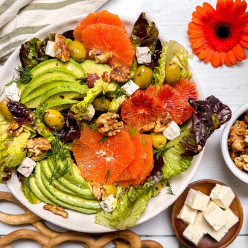 A colorful salad with avocado slices, orange and blood orange rounds, lettuce, olives, walnuts, and feta on a white plate. A wooden spoon, feta cubes, walnuts, and a bright orange flower surround the plate.