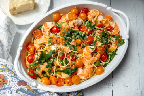 An oval white dish filled with spaghetti, shrimp, halved cherry tomatoes, and chopped parsley. A piece of cheese sits on a plate nearby, with a colorful patterned cloth to the side on a white wooden table.