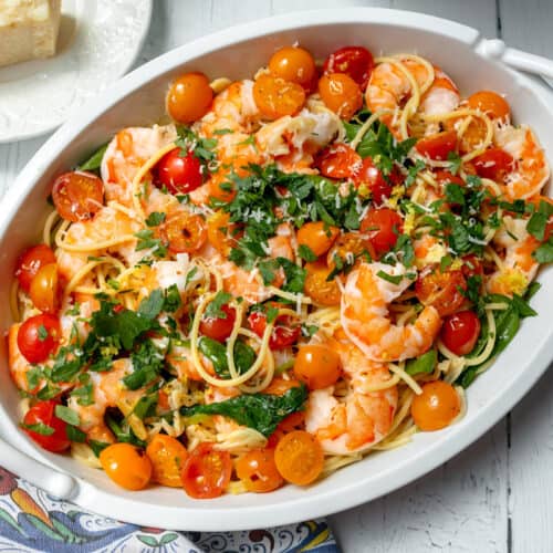 An oval white dish filled with spaghetti, shrimp, halved cherry tomatoes, and chopped parsley. A piece of cheese sits on a plate nearby, with a colorful patterned cloth to the side on a white wooden table.