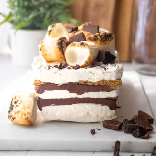A rectangular no-bake s'mores icebox cake topped with toasted marshmallows and chocolate chunks sits on a white marble board, with extra chocolate pieces scattered around. A plant and jars are in the blurred background.