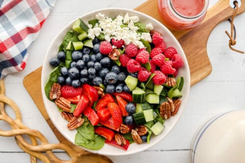A vibrant spinach strawberry salad featuring blueberries, raspberries, cucumber, pecans, and crumbled cheese is served in a bowl with a jar of pink dressing on the side, all set on a rustic wooden board.