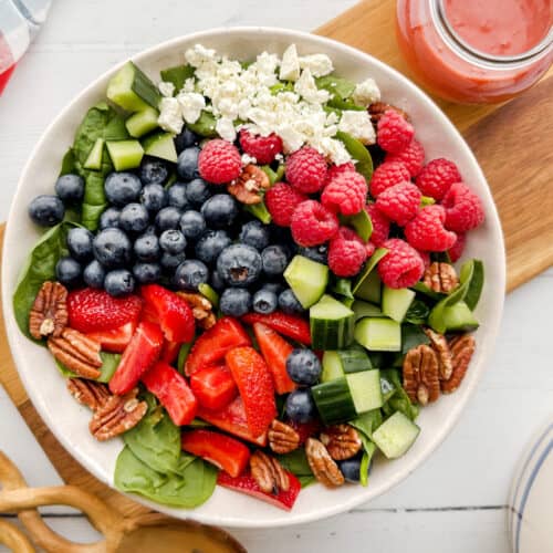 A vibrant spinach strawberry salad featuring blueberries, raspberries, cucumber, pecans, and crumbled cheese is served in a bowl with a jar of pink dressing on the side, all set on a rustic wooden board.