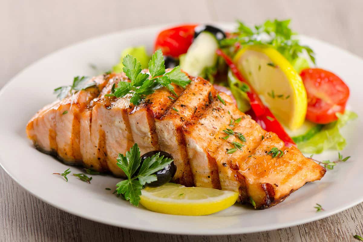 Honey glazed salmon fillet garnished with parsley, served with lemon slices, black olives, and a fresh salad of tomatoes, cucumber, and lettuce on a white plate.