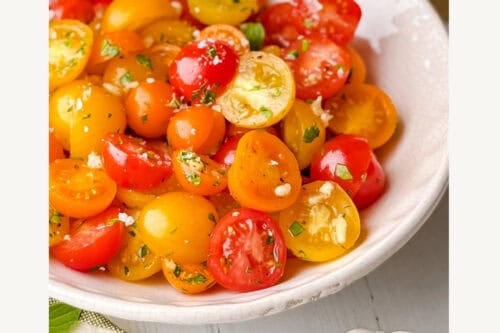 A bowl of halved red and yellow cherry tomatoes mixed with herbs sits on a white table, with a striped napkin, garlic, and a wooden container in the background. Text reads: "5 Minute Marinated Tomatoes. Get the secrets!.
