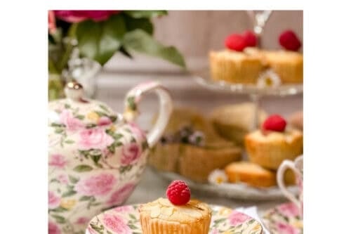 A Mini Bakewell Tart topped with a raspberry rests on a floral plate and napkin. In the background, a teapot, teacup, and more Mini Bakewell Tarts create a charming afternoon tea scene. Text reads "Afternoon Tea Mini Bakewell Tarts.