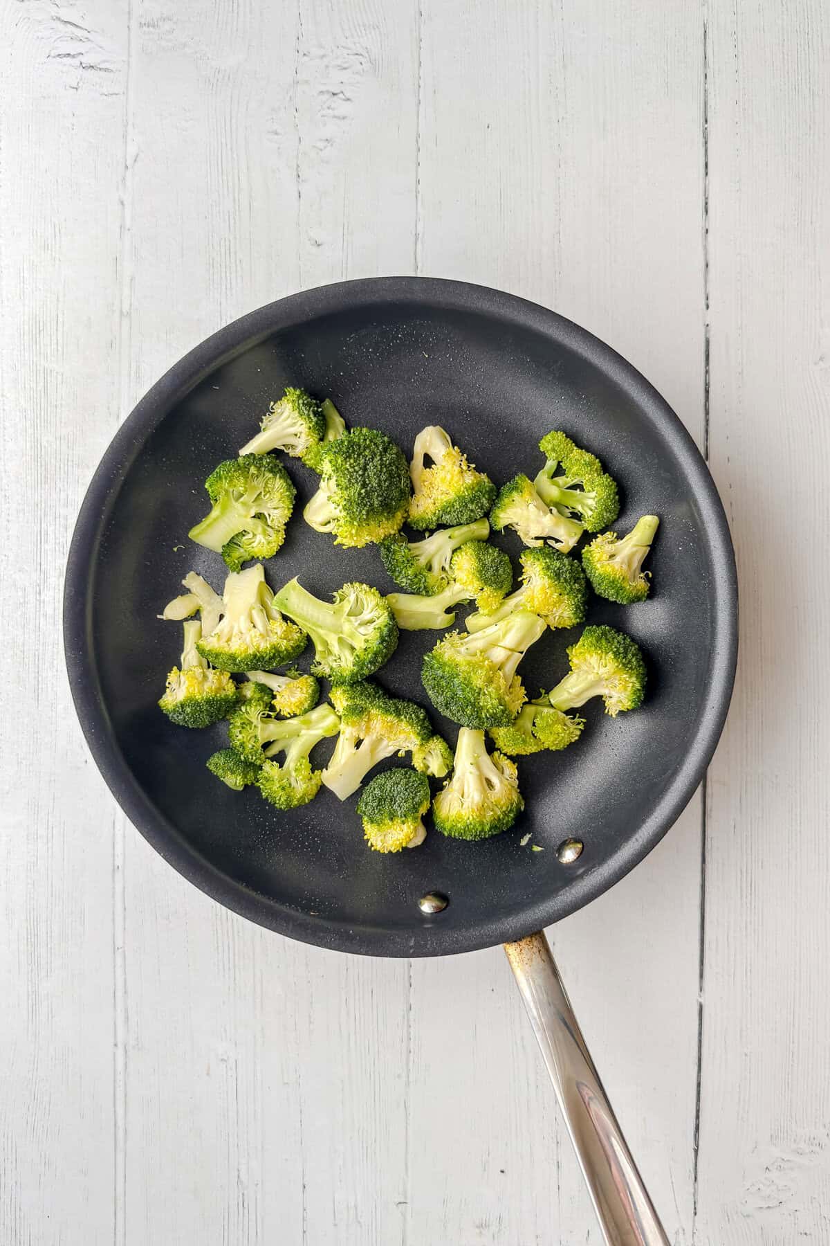 A nonstick frying pan with cooked broccoli florets sits on a white wooden surface, ready to be paired with vibrant Asian Coleslaw for a fresh and flavorful meal.