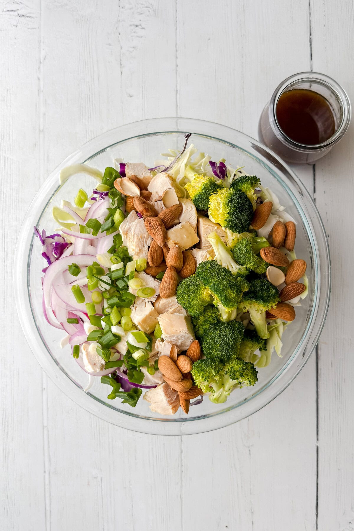 A glass bowl filled with Asian Coleslaw featuring broccoli, red cabbage, onions, chopped green onions, almonds, and chicken, sits on a white wooden surface next to a small jar of dark dressing.