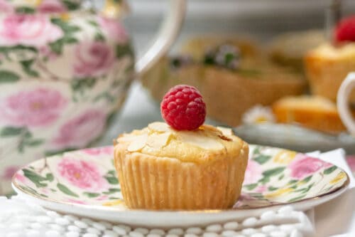 A bakewell tart topped with a raspberry sits on a floral plate, with a floral teapot and more cupcakes in the blurred background. The scene is set on a white, textured cloth with a soft, elegant atmosphere.