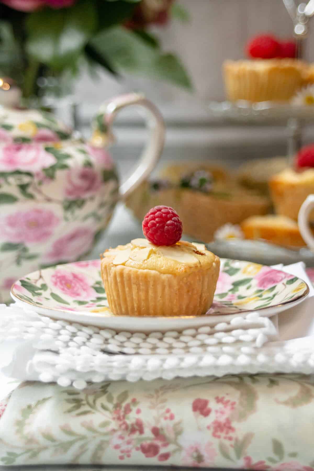 A bakewell tart topped with a raspberry sits on a floral plate, with a floral teapot and more cupcakes in the blurred background. The scene is set on a white, textured cloth with a soft, elegant atmosphere.