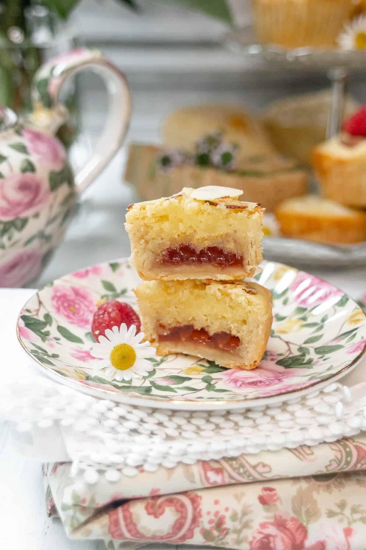 One split almond-topped tarts with a visible raspberry jam filling are stacked on a floral plate, next to a daisy flower. In the background, there is a floral teapot and more baked goods.
