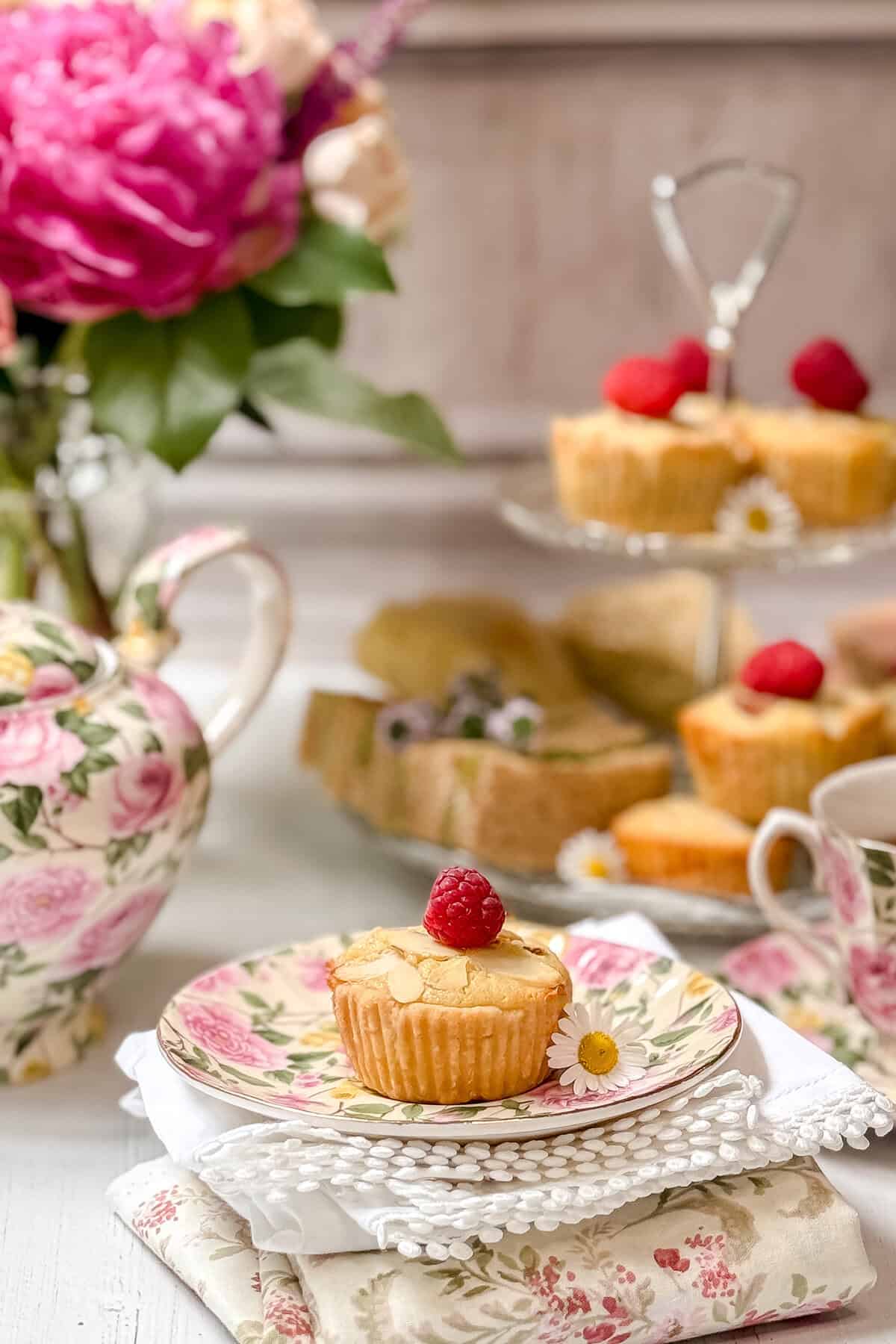 A floral teapot and teacup set beside a plate with a single almond cupcake topped with a raspberry, daisies, and more cupcakes on a tiered stand. Pink flowers decorate the soft, elegant tea party setting.