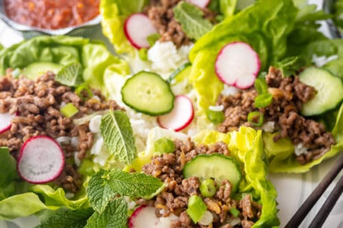 A plate of lettuce wraps filled with seasoned ground meat, sliced radishes, cucumber, green onions, and fresh mint leaves, served with a small dish of red sauce and a side of rice.