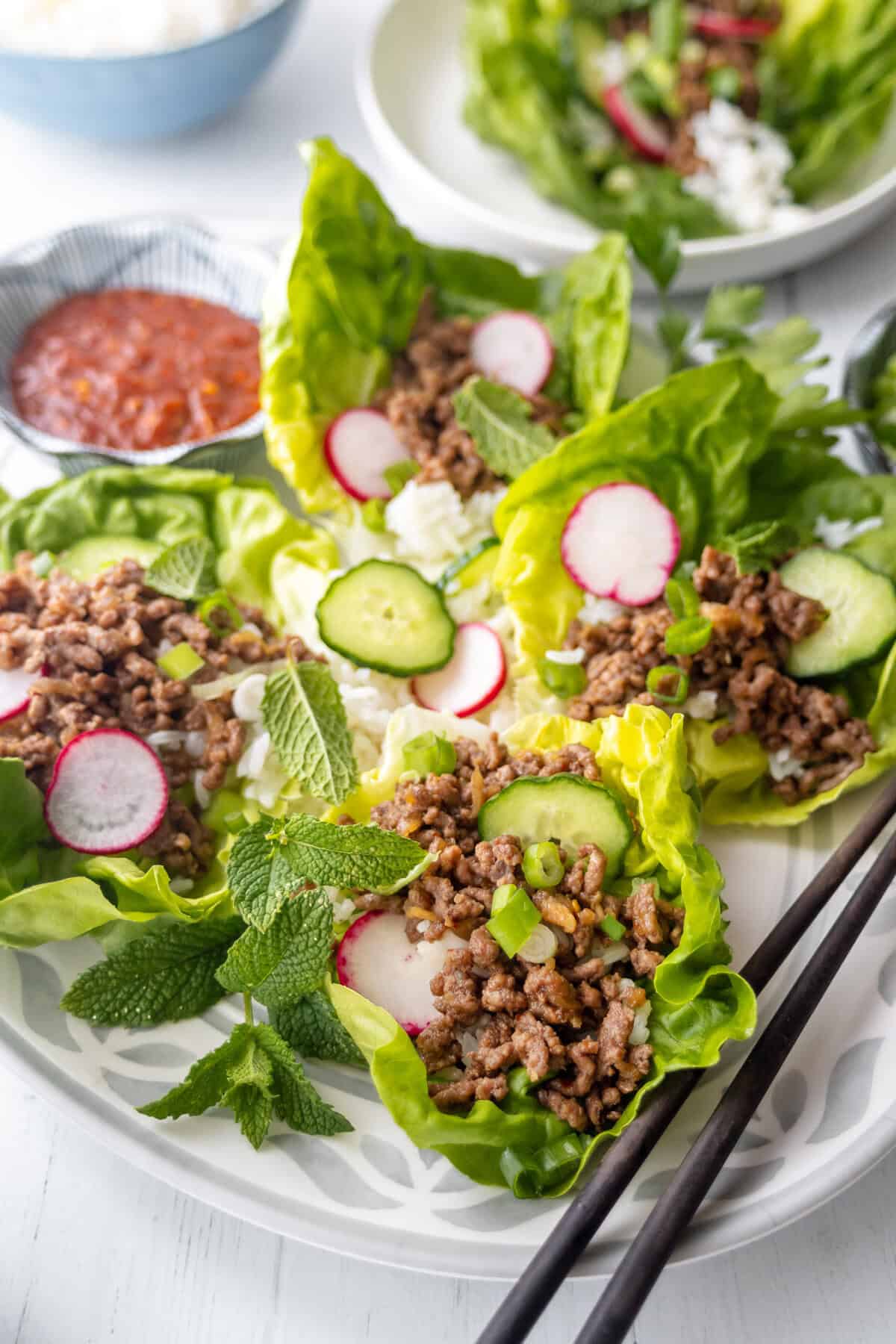 A plate of lettuce wraps filled with seasoned ground meat, sliced radishes, cucumber, green onions, and fresh mint leaves, served with a small dish of red sauce and a side of rice.