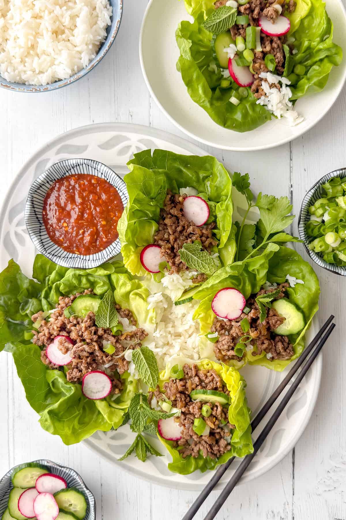 A plate of lettuce wraps filled with Korean beef, rice, sliced radishes, cucumbers, herbs, and green onions. Small bowls of sweet chili sauce, steamed rice, and green onions are on the side. Chopsticks rest nearby.