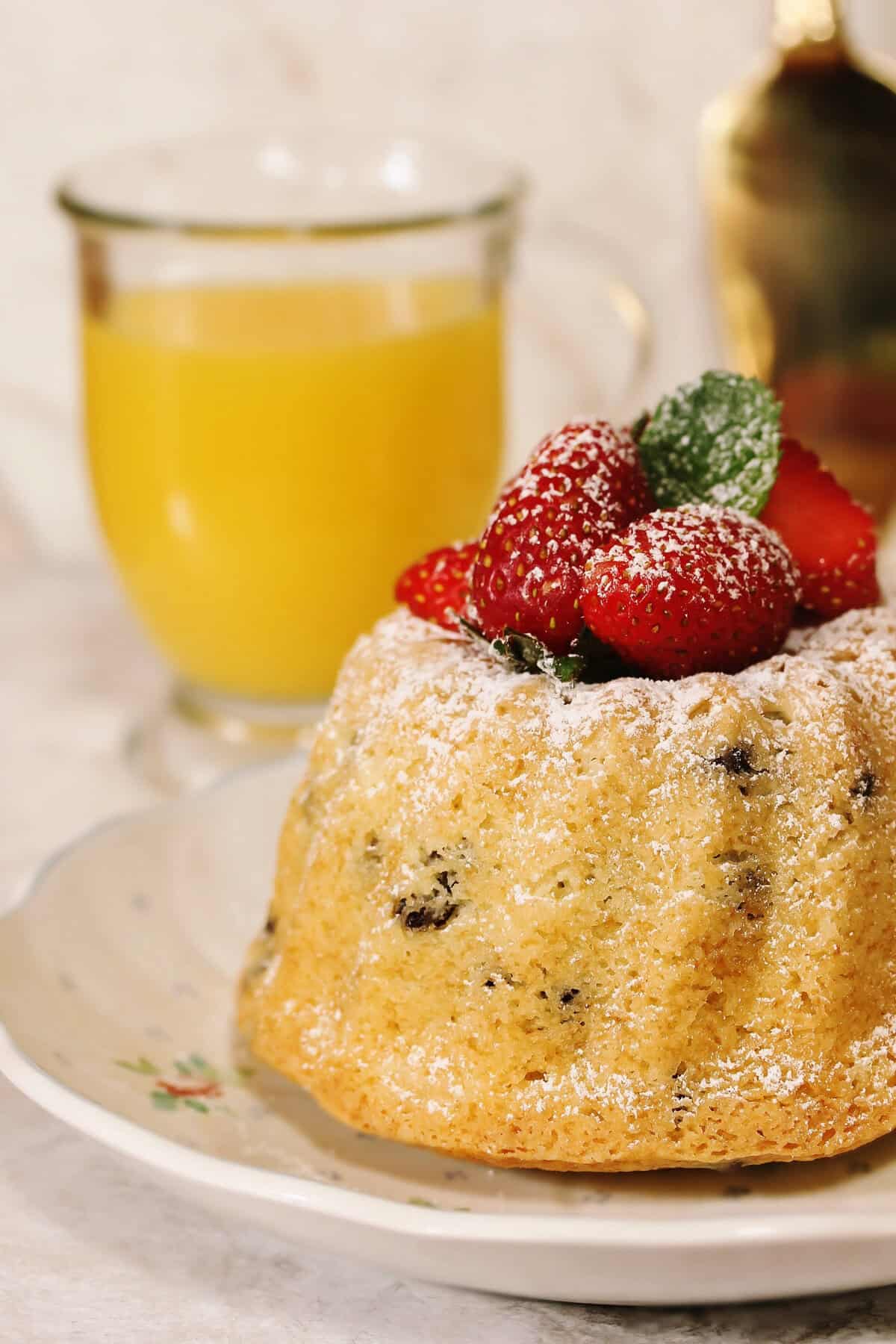 A small bundt cake dusted with powdered sugar, topped with fresh strawberries and mint, sits on a floral plate. A glass mug of orange juice is in the background.