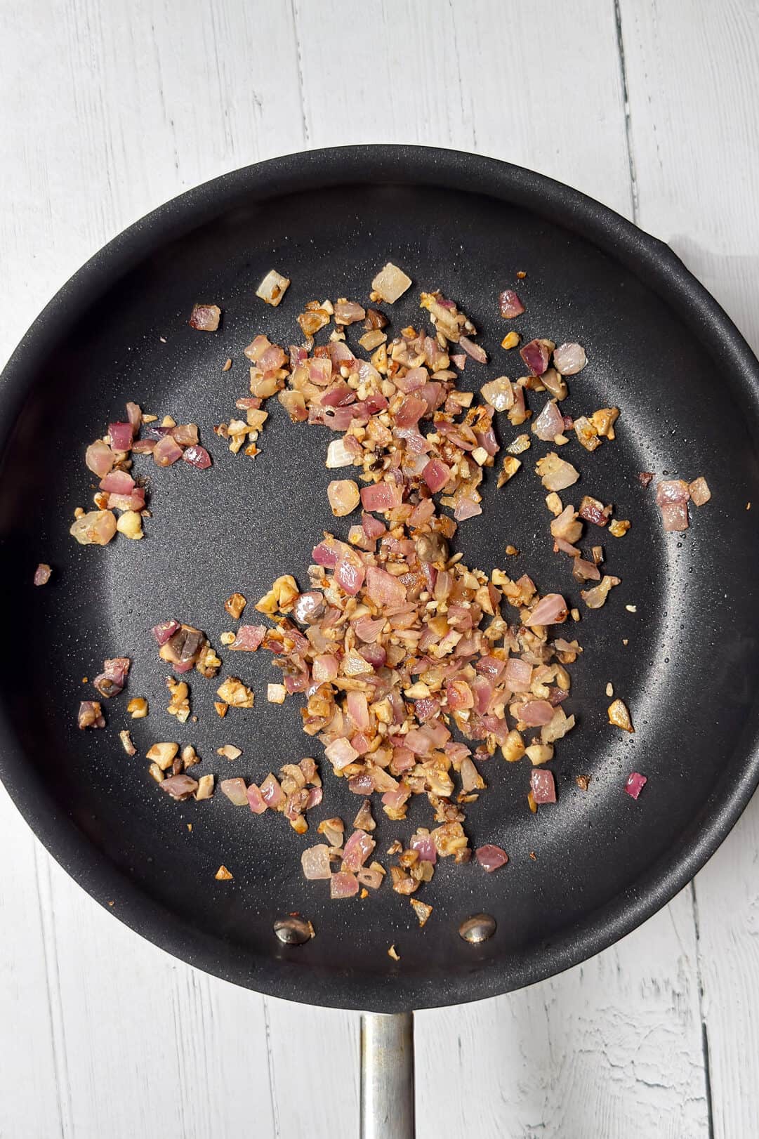 A black frying pan with diced red onions and minced garlic saut&eacute;ing on a white wooden surface.