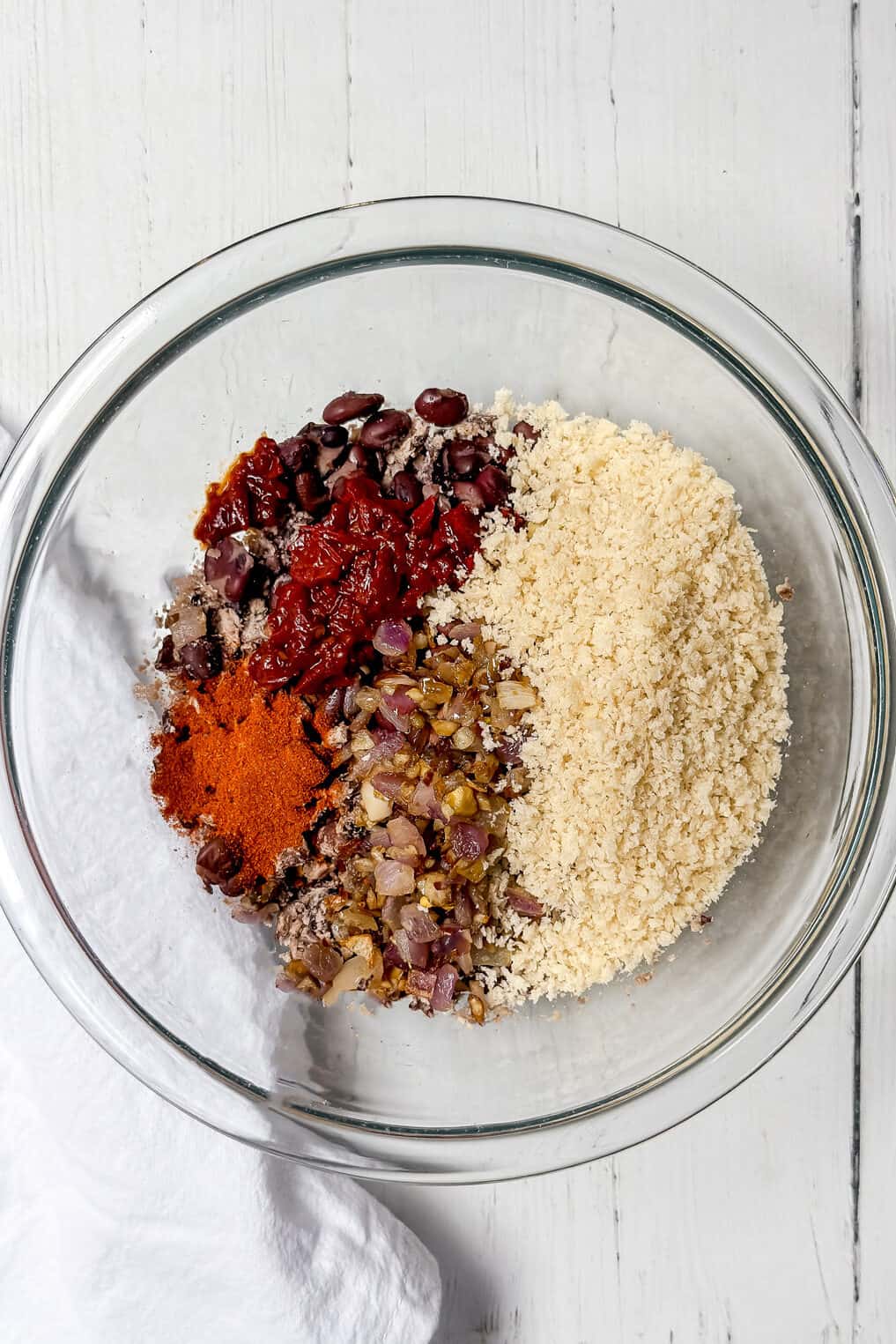 A glass bowl on a white surface containing separate piles of kidney beans, sun-dried tomatoes, almond flour, saut&eacute;ed onions, and paprika. A white cloth is partially visible on the left side.