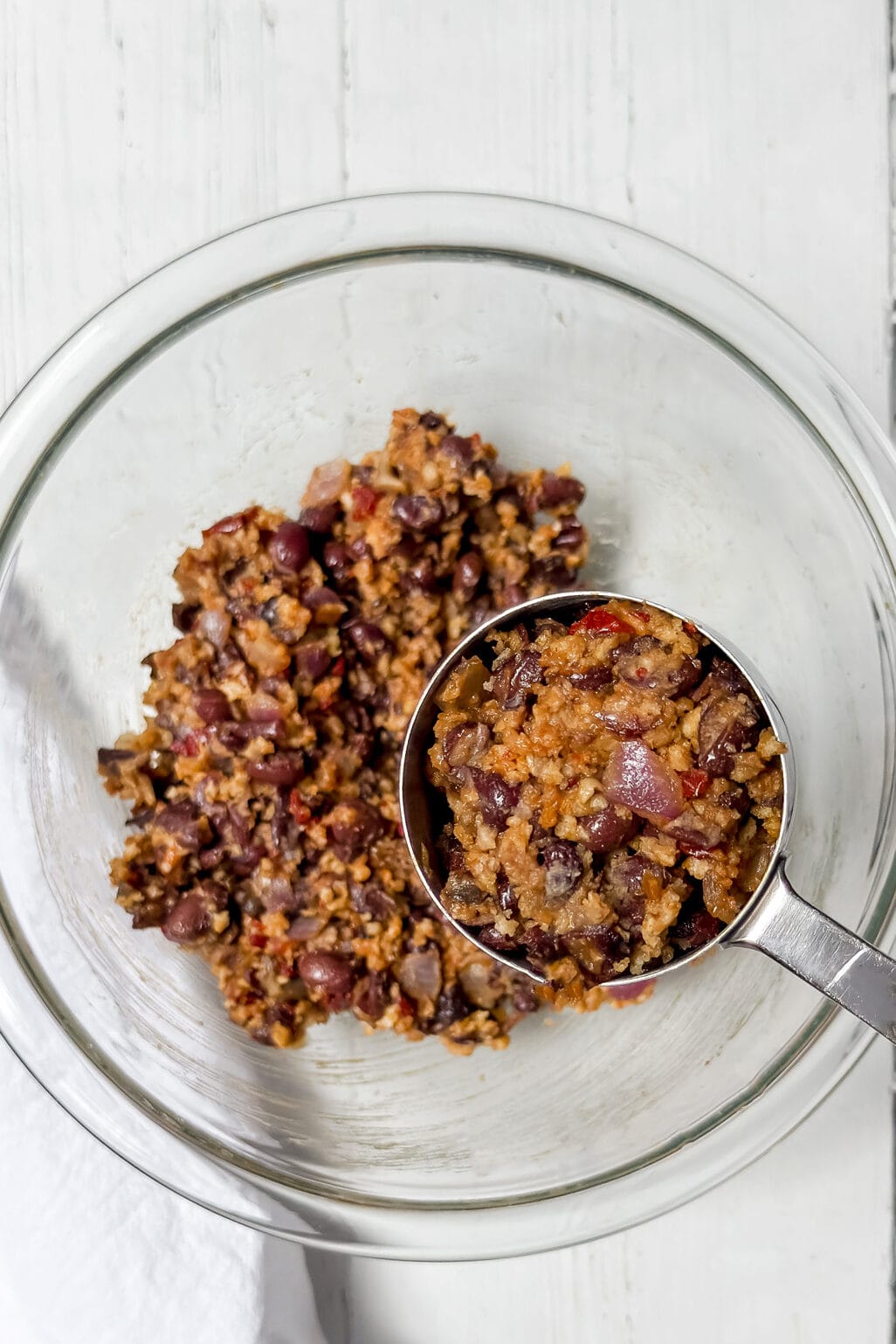 A metal measuring cup scoops a mixture of beans, grains, and vegetables from a clear glass bowl on a white wooden surface.