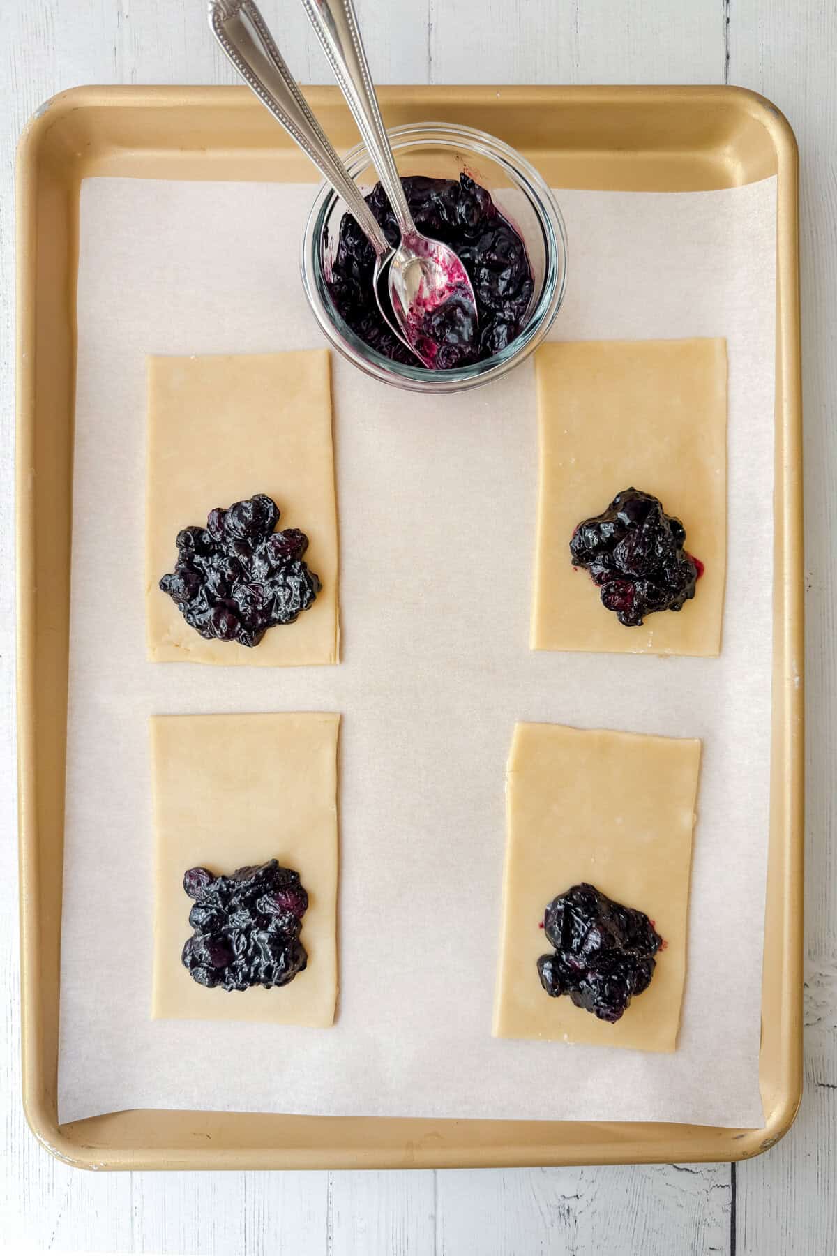 A baking tray lined with parchment holds four rectangular blueberry hand pies, each topped with a spoonful of blueberry filling. A small glass bowl with more filling and two spoons sits at the top of the tray.