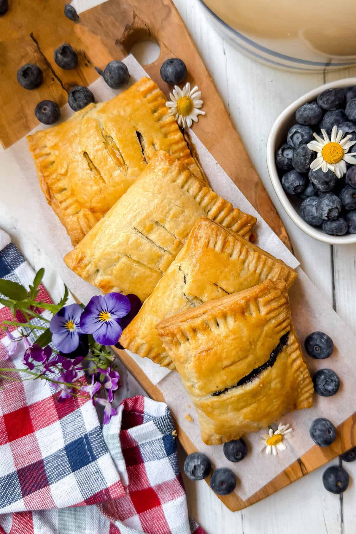 Three golden-brown blueberry hand pies with rich blueberry filling rest on parchment atop a wooden board, surrounded by fresh blueberries, small flowers, and a red-and-blue checkered cloth.