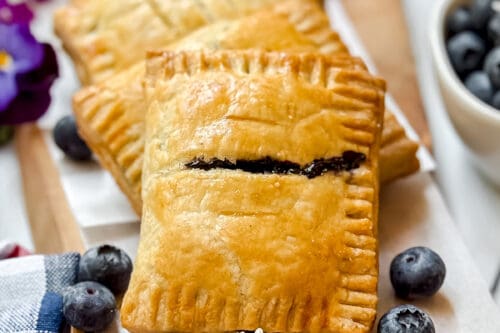Four golden-brown blueberry hand pies with a sweet filling are arranged on a wooden board, decorated with fresh blueberries and small daisies, next to a bowl of blueberries and a colorful napkin.