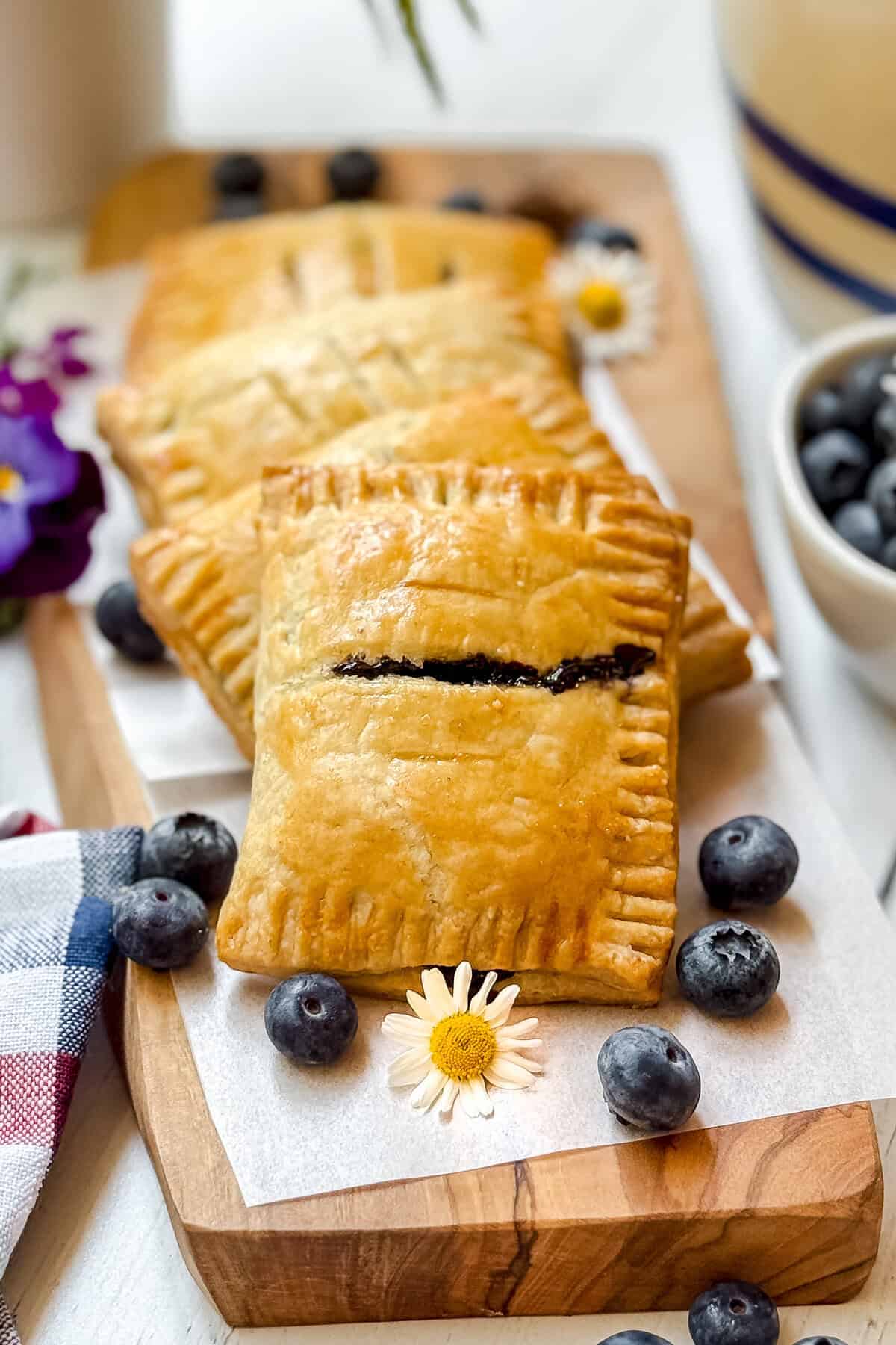 Four golden-brown blueberry hand pies with a sweet filling are arranged on a wooden board, decorated with fresh blueberries and small daisies, next to a bowl of blueberries and a colorful napkin.