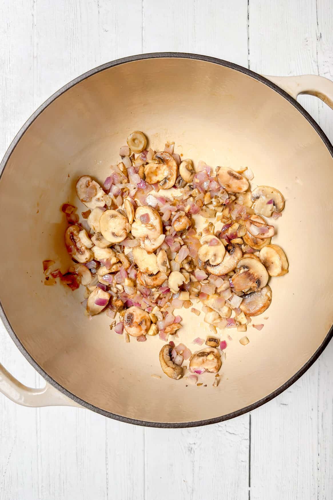 Overhead view of a large cream-colored pot on a white wooden surface, with sliced mushrooms and chopped red onions saut&eacute;ing inside-the perfect start for a delicious One Pot Pasta.