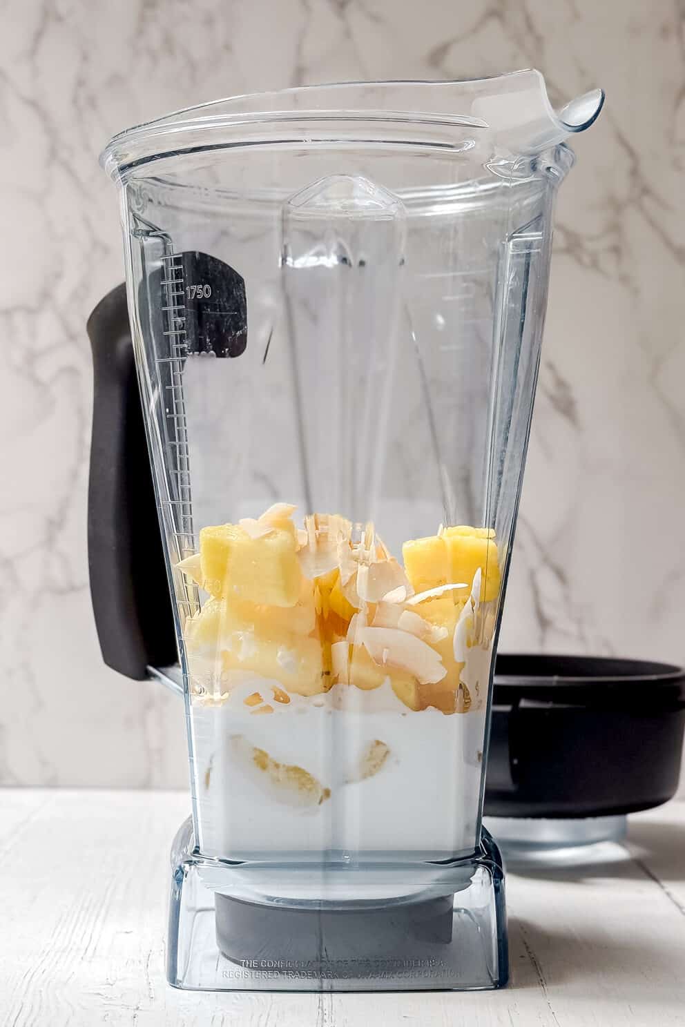 A clear blender jar filled with chunks of pineapple, banana, coconut flakes, and white liquid sits on a white wooden surface with a marble-patterned background—perfect for making a fresh pineapple smoothie. The blender lid rests nearby.