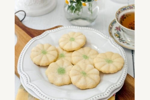 Plate of round, flower-shaped Girl Scout Cookies with green centers on a white plate, a cup of tea, and a small vase of flowers in the background. Text reads: "Girl Scouts Cookies Original Recipe. Get the recipe.