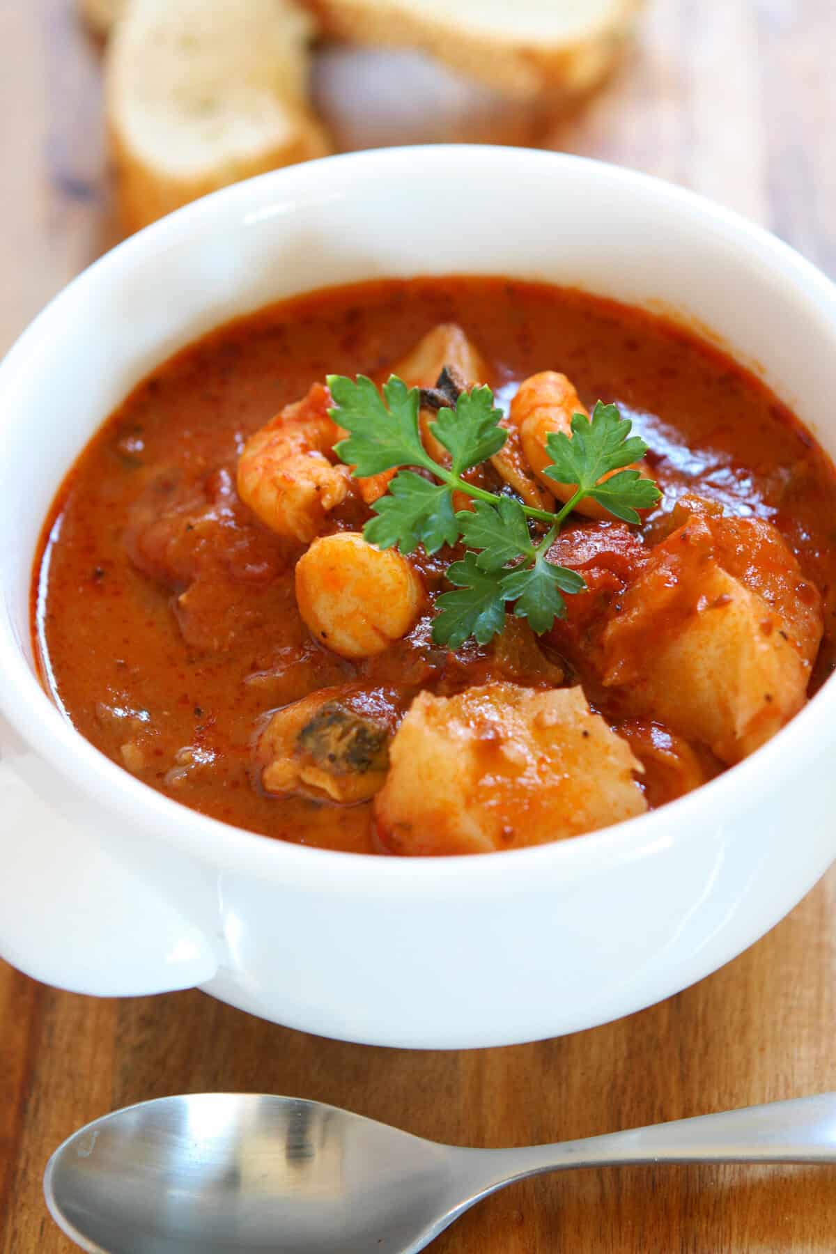 A white bowl of chunky seafood stew with tomato-based broth, garnished with fresh parsley, sits on a wooden surface near a silver spoon and slices of bread in the background.