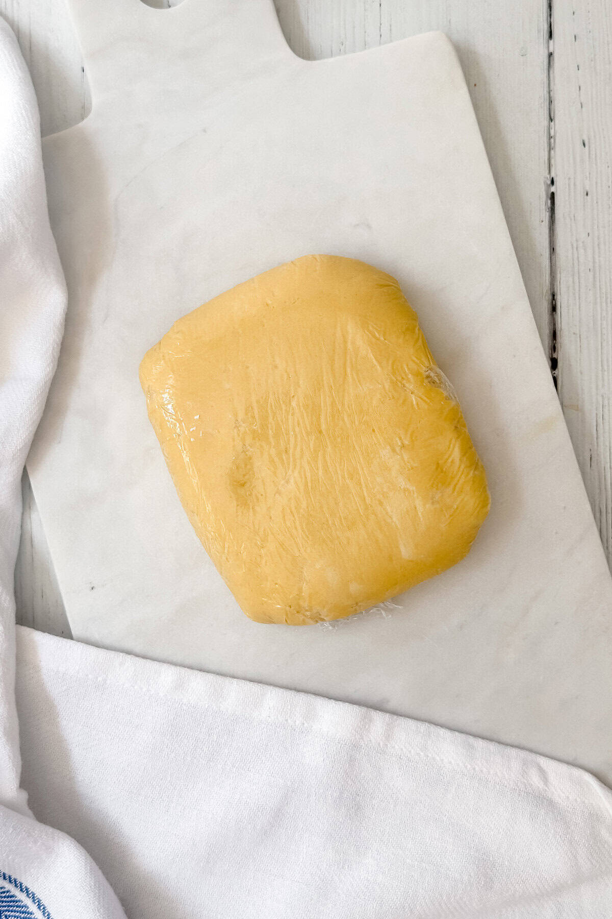 A block of yellow dough wrapped in plastic wrap sits on a white marble cutting board, ready to be shaped into homemade treats inspired by Girl Scout Cookies, with white cloths nearby on a light wooden surface.