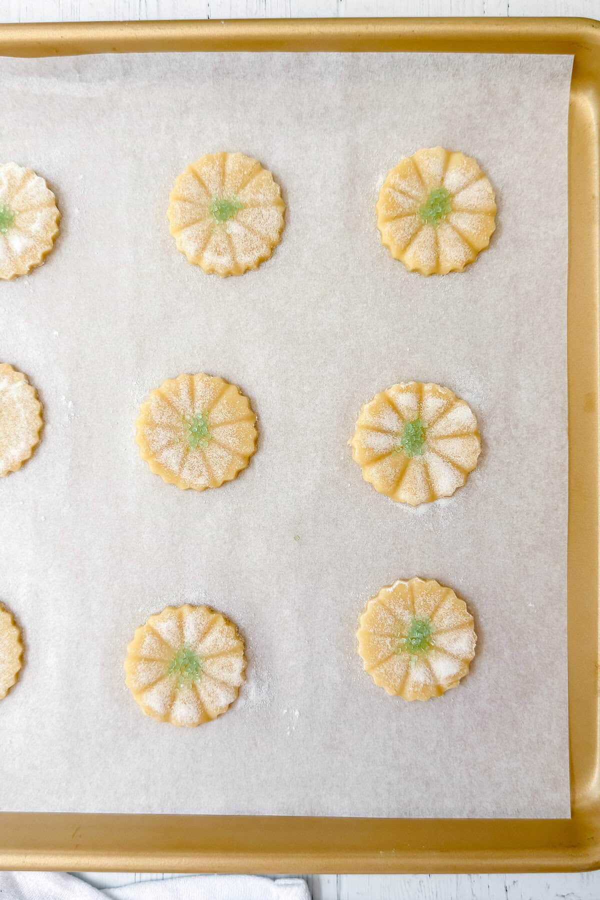 A baking sheet with nine uncooked, flower-shaped sugar cookies-reminiscent of classic Girl Scout Cookies-topped with green sugar in the center and arranged evenly on parchment paper.
