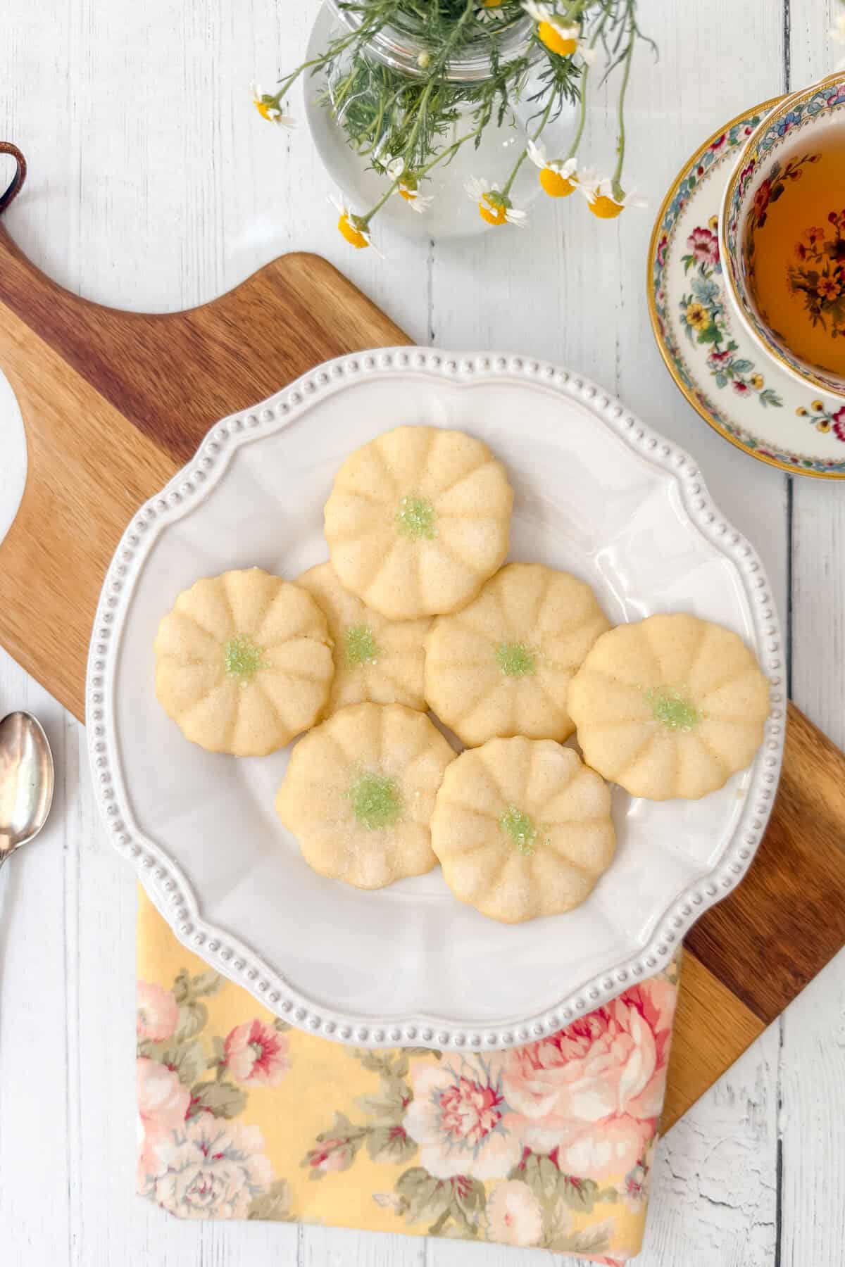 A white plate with six flower-shaped sugar cookies, reminiscent of Girl Scout Cookies and topped with green sprinkles, sits on a wooden board. Nearby are a spoon, floral napkin, cup of tea, and a vase with small yellow flowers.